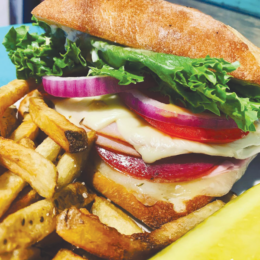 A close up shot of a sandwich made from a ciabatta roll, bright vegetables, ham and cheese, with a side of fries