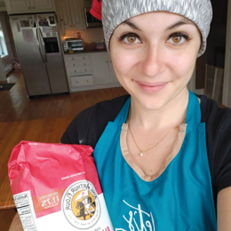 A photo of a young woman standing in her kitchen, holding a bag of King Arthur Flour.