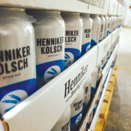 Upclose photo of cases of Henniker Brewery Henniker Kolsche cans, with a minimalist blue and white label