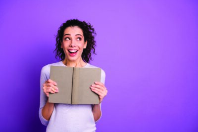curly wavy charming cute nice lady impressed after reading, her paper book excited ecstatic smiling toothily isolated over purple pastel color background