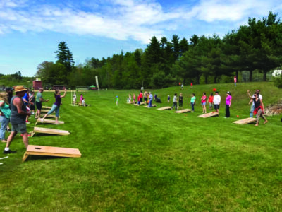 green field with tree in background, lines of people playing cornhole