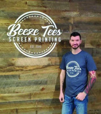 man in t-shirt standing against wooden wall with Beeze Tees logo painted on it