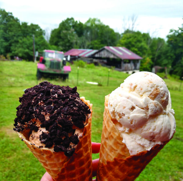 Hand holding up two ice cream cones in front of farm field, truck and barn in background