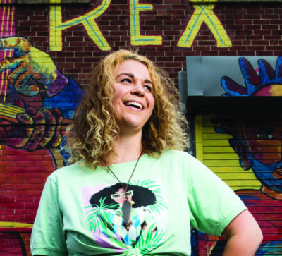 young curly haired blonde woman in t-shirt, looking left and smiling, standing in front of mural on brick wall
