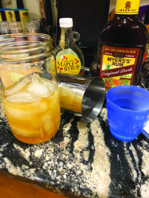 mason jar with ice and cocktail, bottle of rum, bottle of maple syrup, drink mixing cup, sitting on marble counter