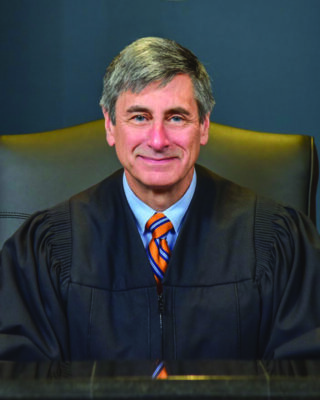 man in judges robe, sitting in chair, wearing blue shirt and orange striped tie