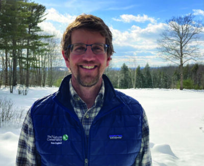 smiling man with glasses, wearing plaid shirt and winter vest, standing in front of snowy woods