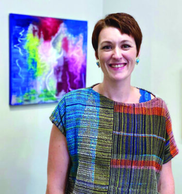 smiling woman wearing colorful woven top, standing in front of painting on a white wall