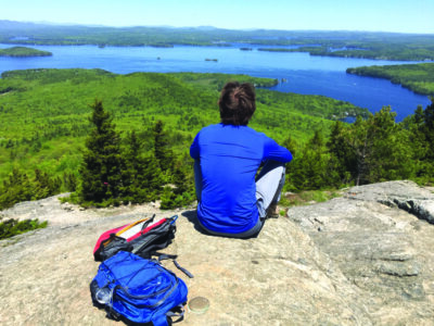 young man sitting on rocky peak, overlooking lake and forest vista on sunny day