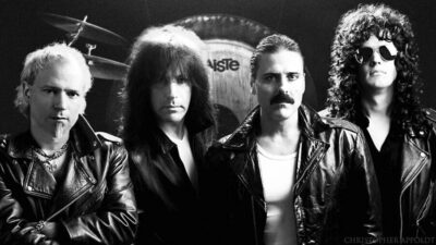 four men wearing leather jackets, standing in line in front of drum set