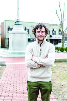 man in sweater and glasses, standing in front of outdoor monument
