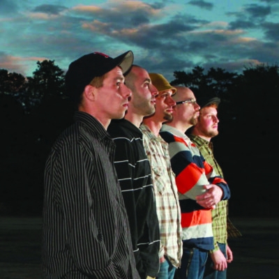 five men standing in line, facing right, in front of trees at dusk
