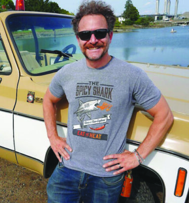 smiling man wearing sunglasses and a t-shirt that reads "The Spicy Shark", leaning on old car in front of body of water