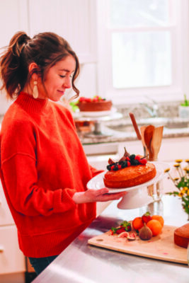 young woman in bright kitchen holding cake platter containing cake with berries on top