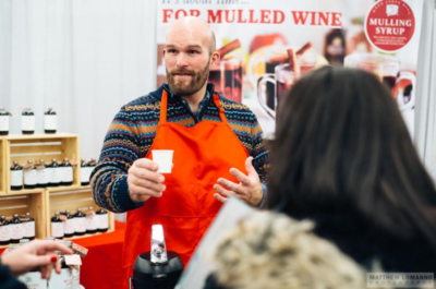 man standing in front of poster advertising mulled wine, speaking with event attendees and handing out samples
