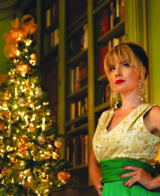 young woman in evening dress standing in room with built-in bookshelf and lit Christmas tree