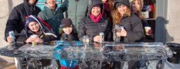 A large family of all ages posing behind a table carved out of ice, all holding a warm drink.