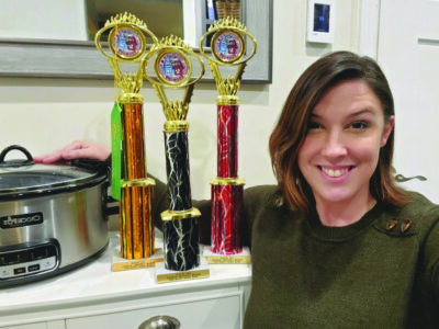 woman in kitchen posing with 3 trophies and a crockpot