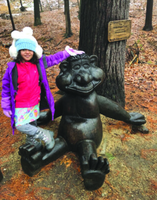 young girl dressed for winter standing with troll sculpture installed along walking trails behind Stark Park
