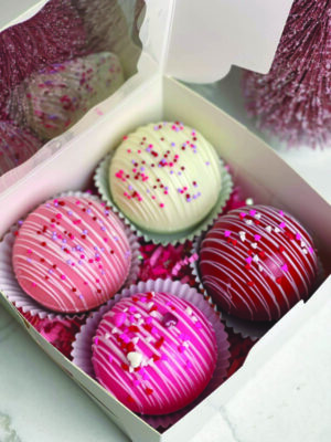4 pink and white cocoa bombs decorated with round and heart-shaped sprinkles, in box