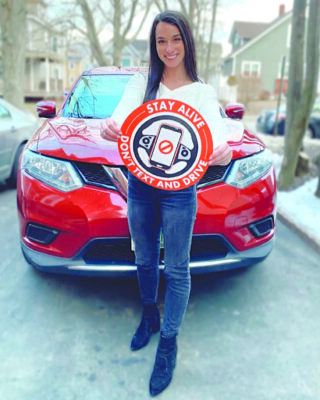 young woman standing in front of car and holding sign warning about texting and driving danger