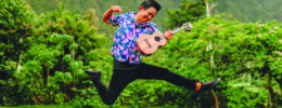 young man leaping and playing ukulele, in field with mountain behind