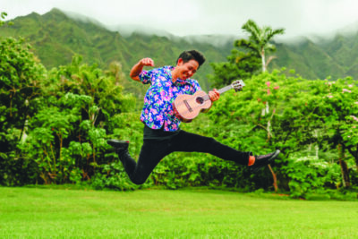 young man leaping and playing ukulele, in field with mountain behind