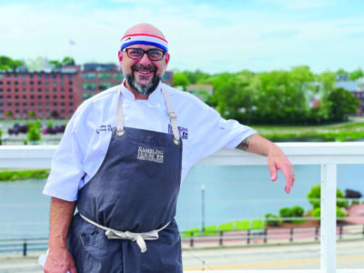 bearded man standing in front of river and skyline, wearing apron, smiling