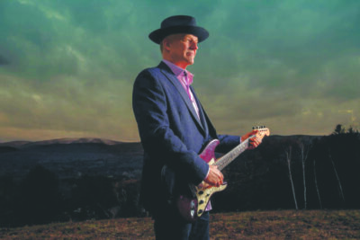 man in suit and brimmed hat holding guitar, posing in front of mountain vista