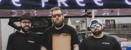 3 men standing under banner, man in the middle holding open pizza box displaying pizza