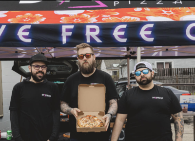 3 men standing under banner, man in the middle holding open pizza box displaying pizza