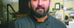 bearded man in dark chef's coat standing in industrial kitchen