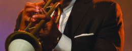 black man wearing suit, playing trombone under stage lights