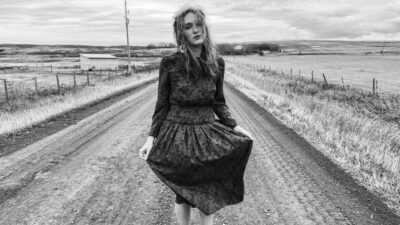black and white image of young woman standing on dirt road, hands on skirt