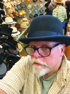 selfie taken from above of man with mustache and chin beard wearing bowler hat, wall of hats on display behind him