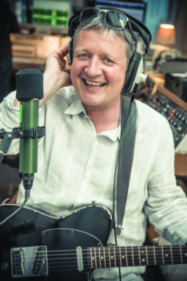 man smiling, sitting in recording studio holding guitar in front of microphone