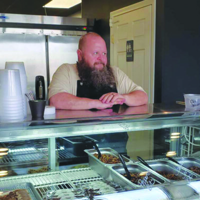bearded man wearing dark apron, standing behind food counter in restaurant