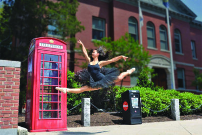 ballerina leaping, outside, on sidewalk in front of brick building and phone booth