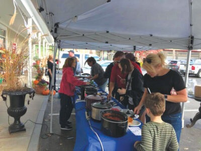 Group of people gathered under a pop up tent, tasting different crock pots of chili - They're not double dipping I swear