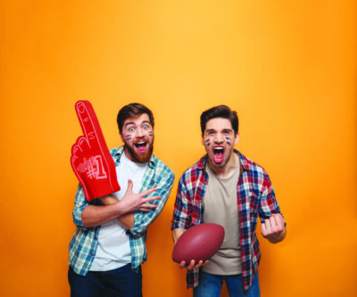 Portrait of a two happy young men holding rugby ball and foam glove while celebrating isolated over yellow background