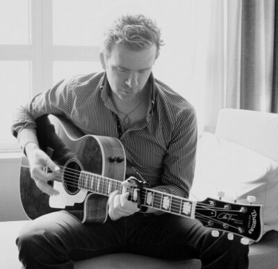 Black and white photo of Chris Trapper, a very rock and roll looking younger man playing an acoustic guitar.