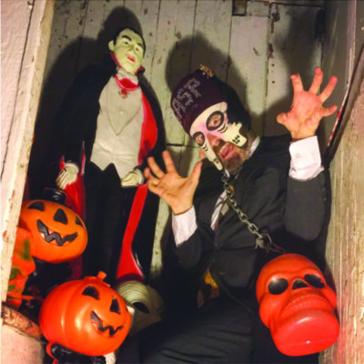 man wearing black suit and partial face mask, posing with vampire dummy and pumpkin shaped trick or treat baskets