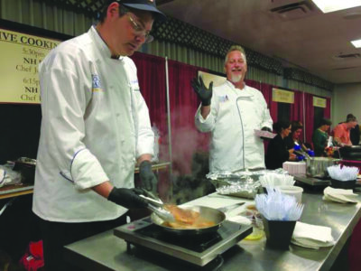 2 men in chef's coats, cooking at table at food expo