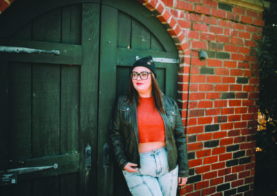 woman in jeans and leather jacket, wearing beanie hat, leaning against door in brick wall