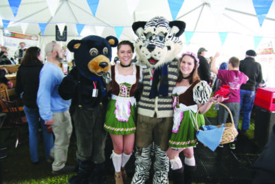 animal mascots, a white tiger and a bear, posing with 2 women dressed in Oktoberfest mini dresses in crowded tent