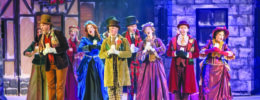 group of actors in colorful Victorian costume on stage singing carols