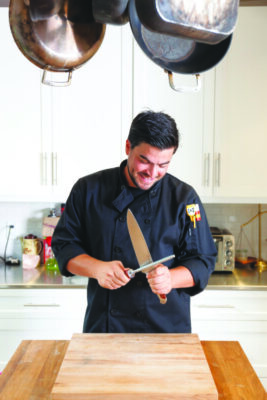 man dressed in black chef jacket, standing in kitchen in front of table, holding knife in eahc hand