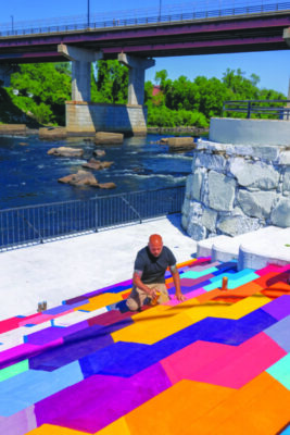 man painting colorful mural on stairs, river behind him
