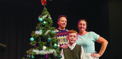 3 actors on stage, woman man and boy child, posing beside Christmas Tree