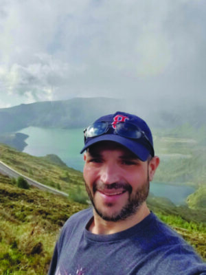 man with beard, wearing baseball cap, smiling, mountain landscape behind